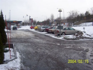 Bild på en parkering med nio bilar i snö, med en väg bakom